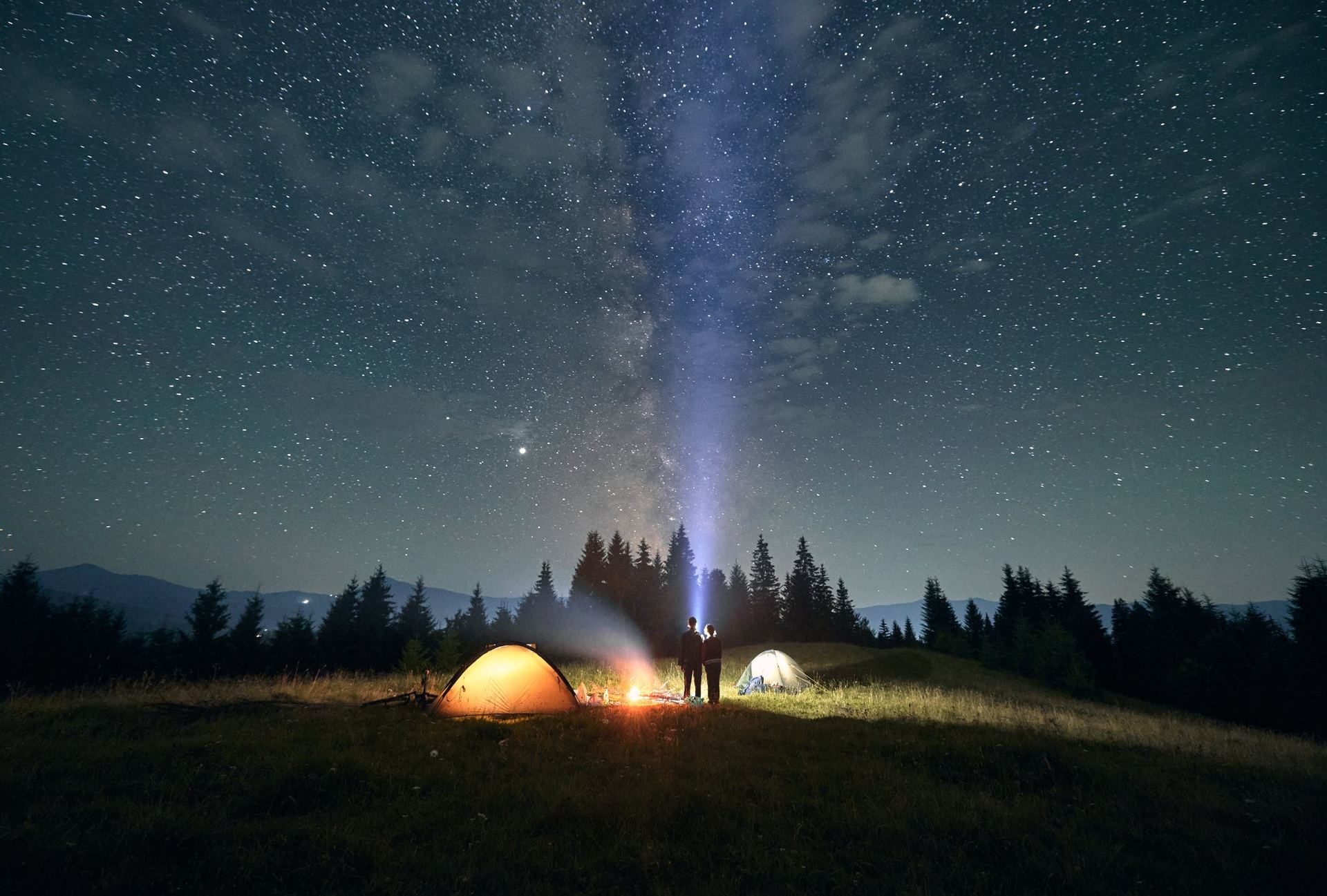Two campers standing by a glowing campfire under the Milky Way
